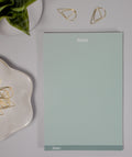 Sage green notes notepad with a green stripe at the bottom, set for the date. The notepad is on a light gray surface surrounded by gold decorative cone-like paper clips at the top and inside a flower-like ceramic bowl next to it. At the top left corner is a snake plant inside a white plant pot.