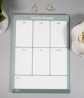 A sage green undated weekly planner notepad with seven white writing sections labeled Mon through Sat/Sun and a notes box, photographed on a light gray surface with small decorative props around it. The decorative props consist of 2 gold cone-like paperclips at the top with more sitting inside a flower-like ceramic bowl next to it and a snake plant inside a white plant pot.