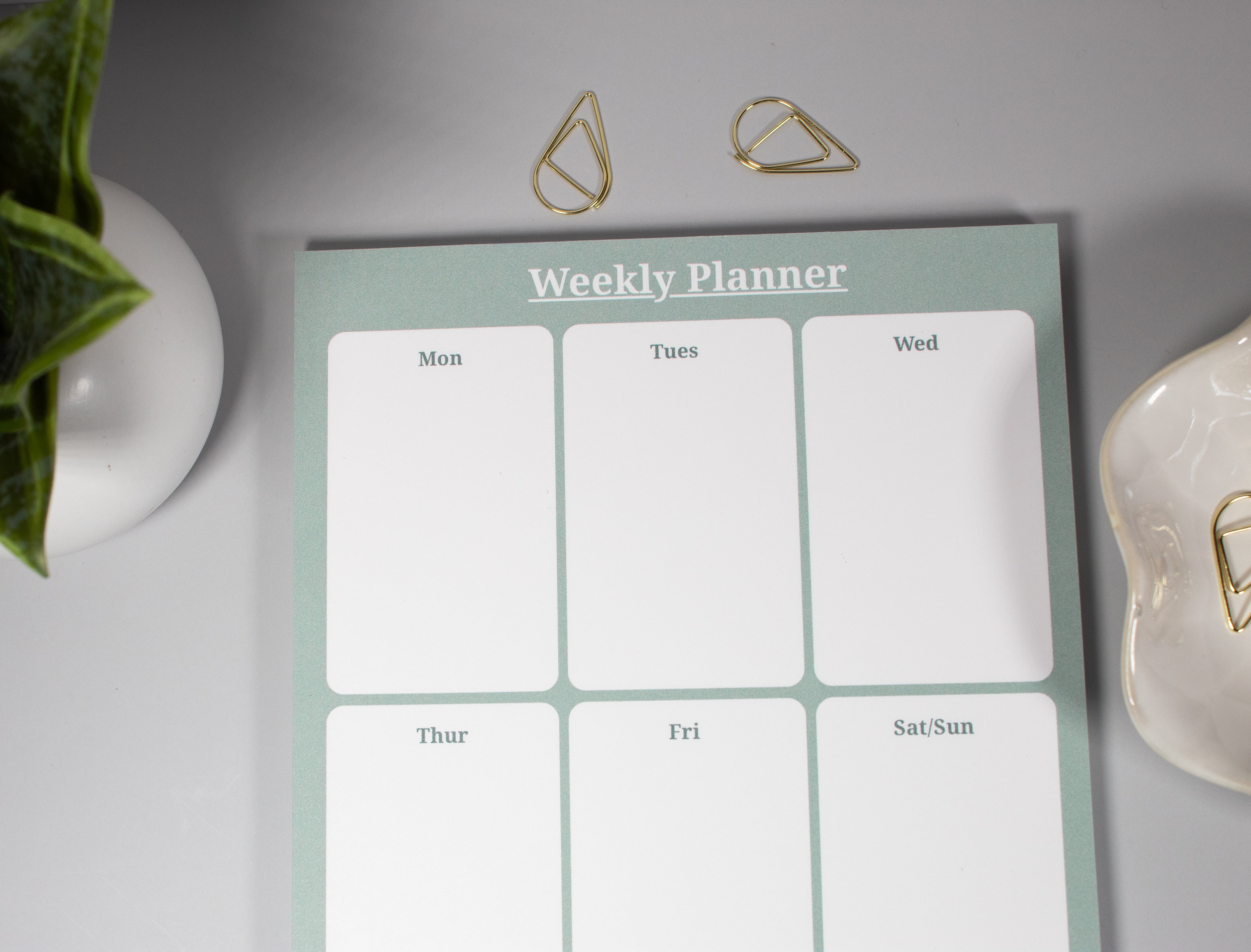A weekly planner notepad with six white daily boxes and a pale sage green border, styled on a light gray surface with gold paper clips, a plant, and a ceramic dish with more gold paperclips.