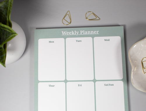 A weekly planner notepad with six white daily boxes and a pale sage green border, styled on a light gray surface with gold paper clips, a plant, and a ceramic dish with more gold paperclips.