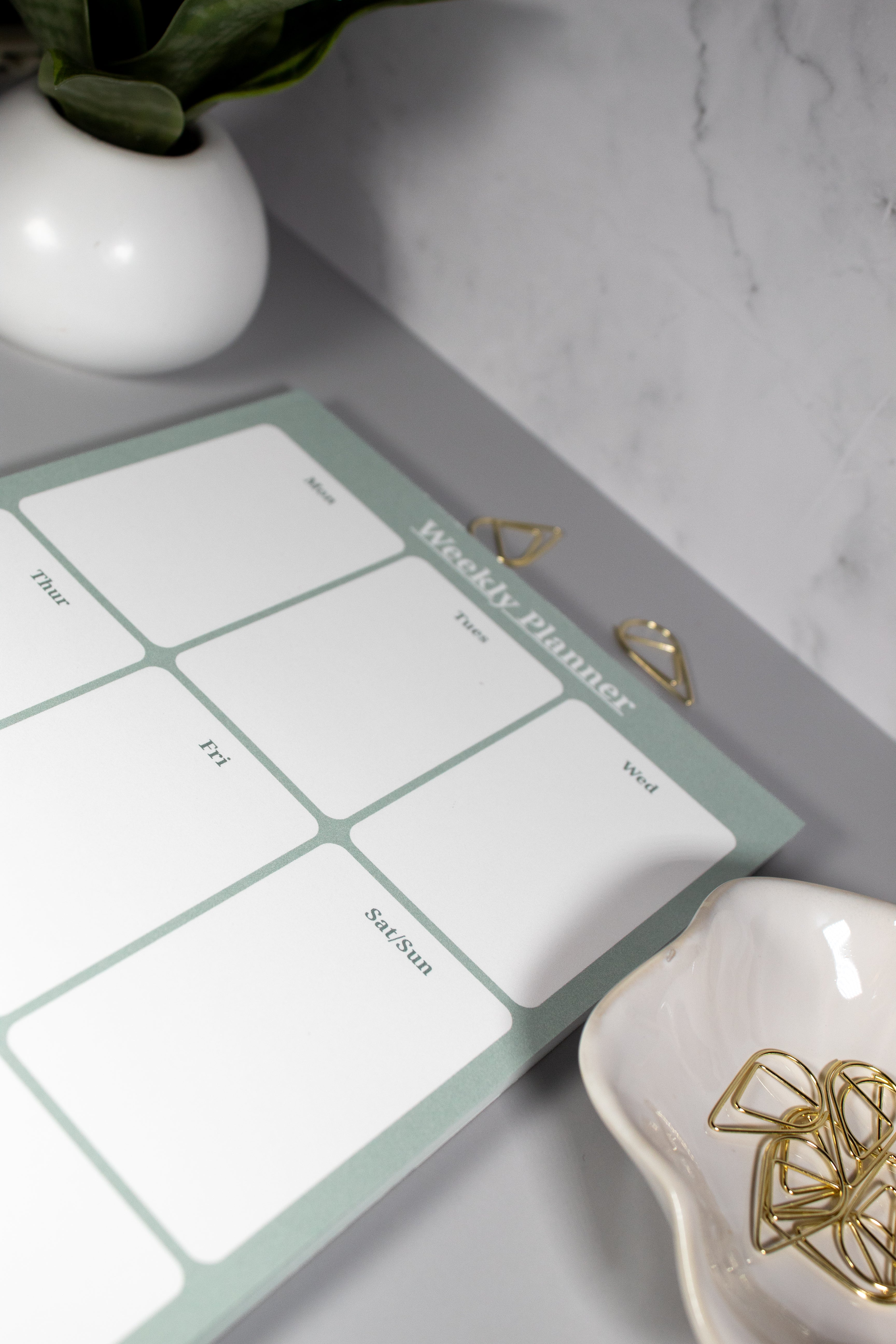 A weekly planner pad with a sage green border and white grid sections, styled on a gray marble surface with decorative paper clips, a white ceramic bowl with more gold paper clips, and a plant in a white vase in the background.