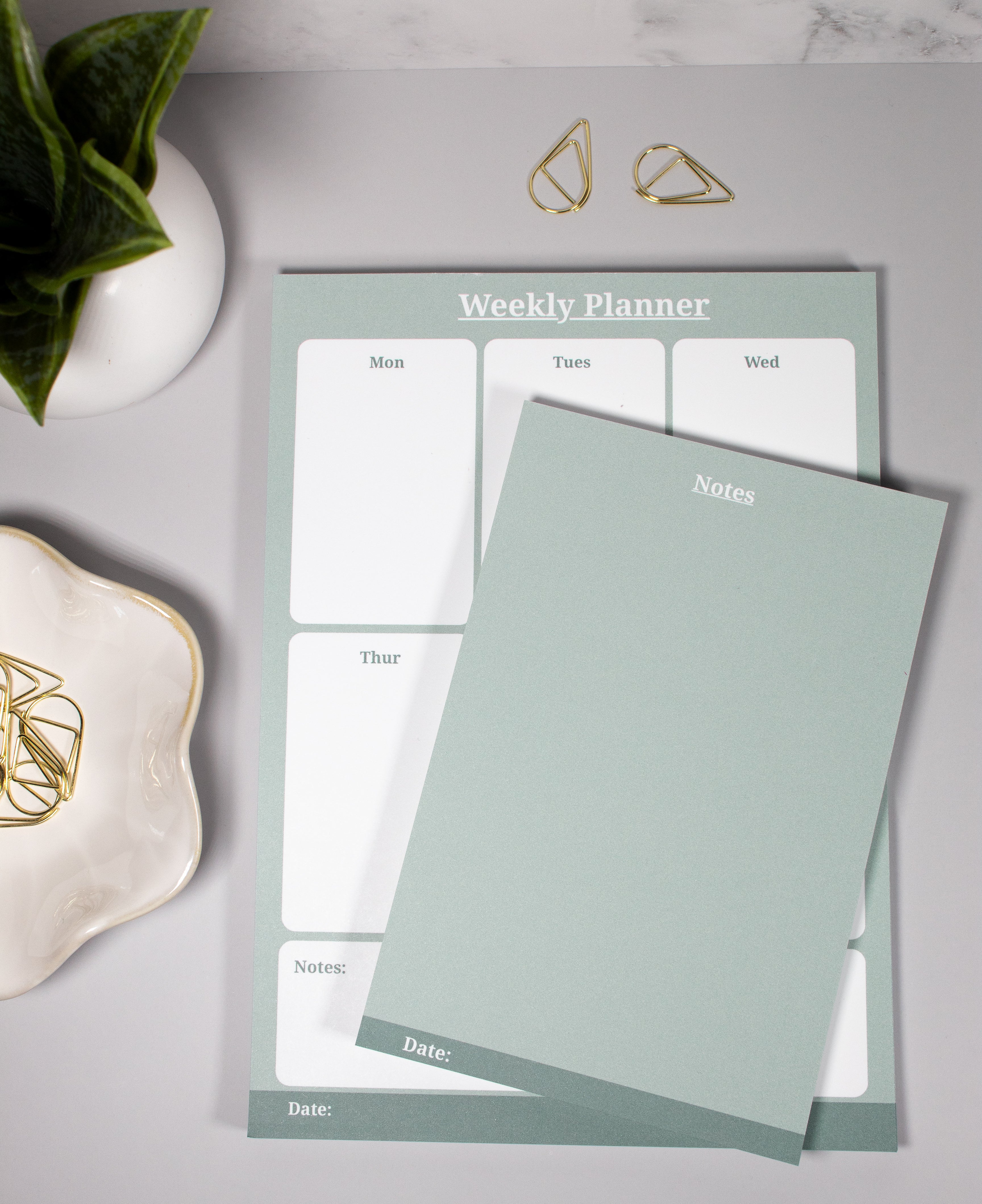 A sage green undated weekly planner pad and matching notes notepad, styled in a flat-lay on a gray surface with gold decorative cone-like paper clips, a small plant, and a flower-like ceramic dish with additional gold paper clips.