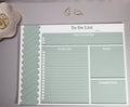 Sage green to-do list notepad with a "Top 3 Priorities" box at the top, the "To-do list" section below to the left. On the right of the to-do list is a "Notes" section and a "Coming Next" section below the "Notes Section". The To-Do list sits on a light gray surface with 2 gold decorative cone-like paper clips at the top with more sitting inside a flower-like ceramic bowl next to it. 