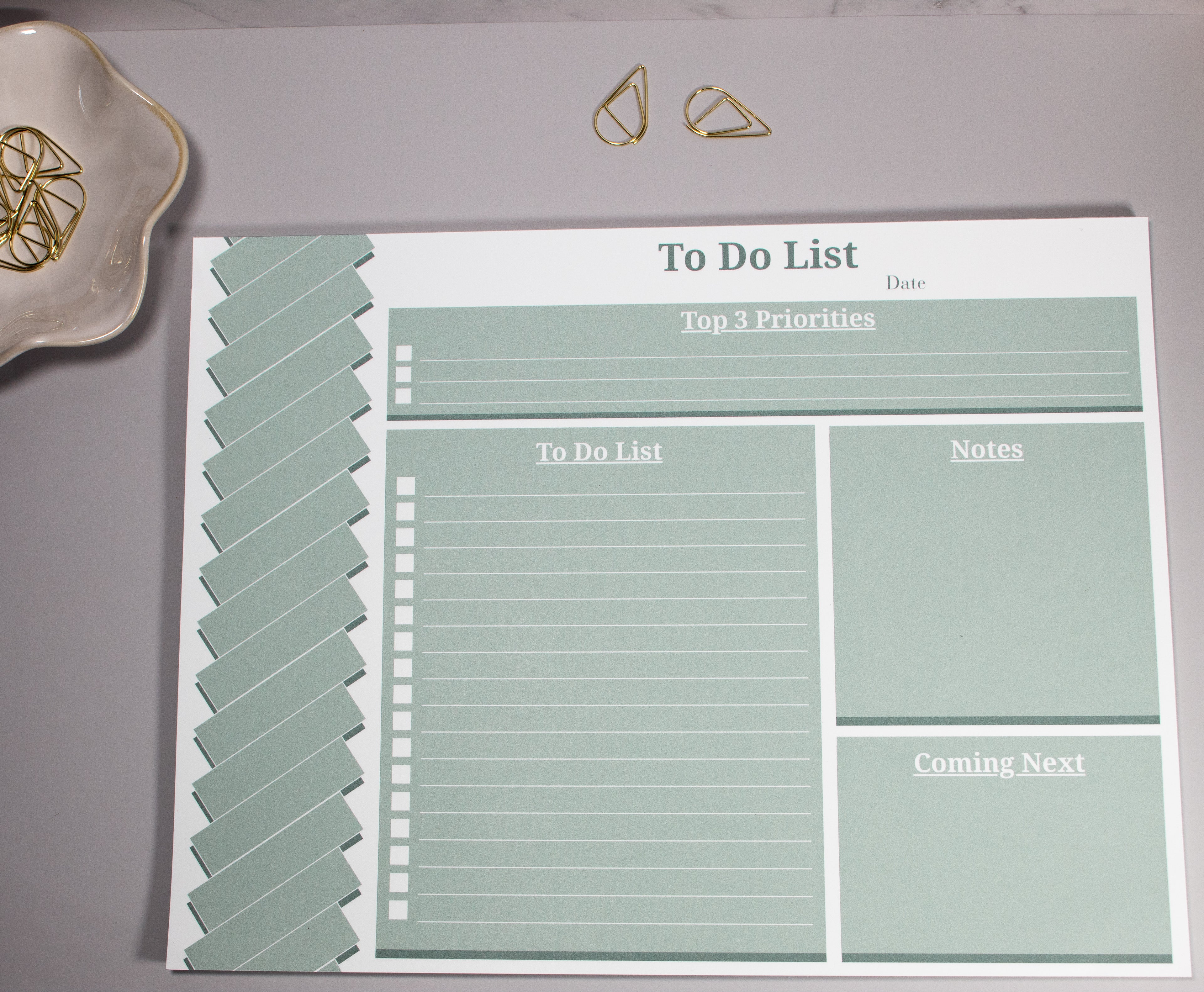 Sage green to-do list notepad with a "Top 3 Priorities" box at the top, the "To-do list" section below to the left. On the right of the to-do list is a "Notes" section and a "Coming Next" section below the "Notes Section". The To-Do list sits on a light gray surface with 2 gold decorative cone-like paper clips at the top with more sitting inside a flower-like ceramic bowl next to it. 