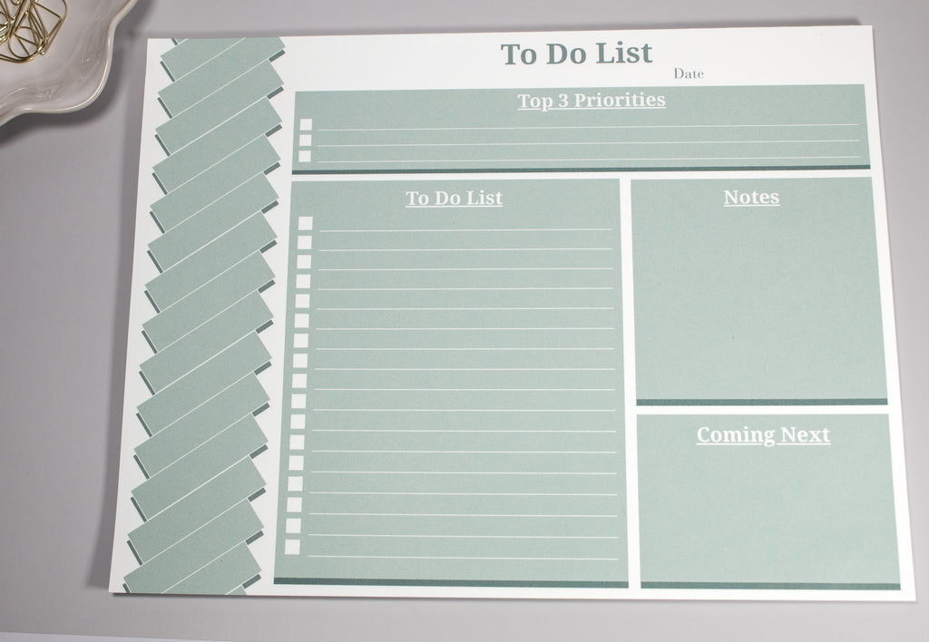 Sage green 8 x 10.5 inch to-do list with a "Top 3 Priorities" box at the top, the "To-do list" section below to the left. On the right of the to-do list is a "Notes" section and a "Coming Next" section below the "Notes Section". There are gold cone-like paperclips at the top sitting inside a flower-like ceramic bowl next to it. 