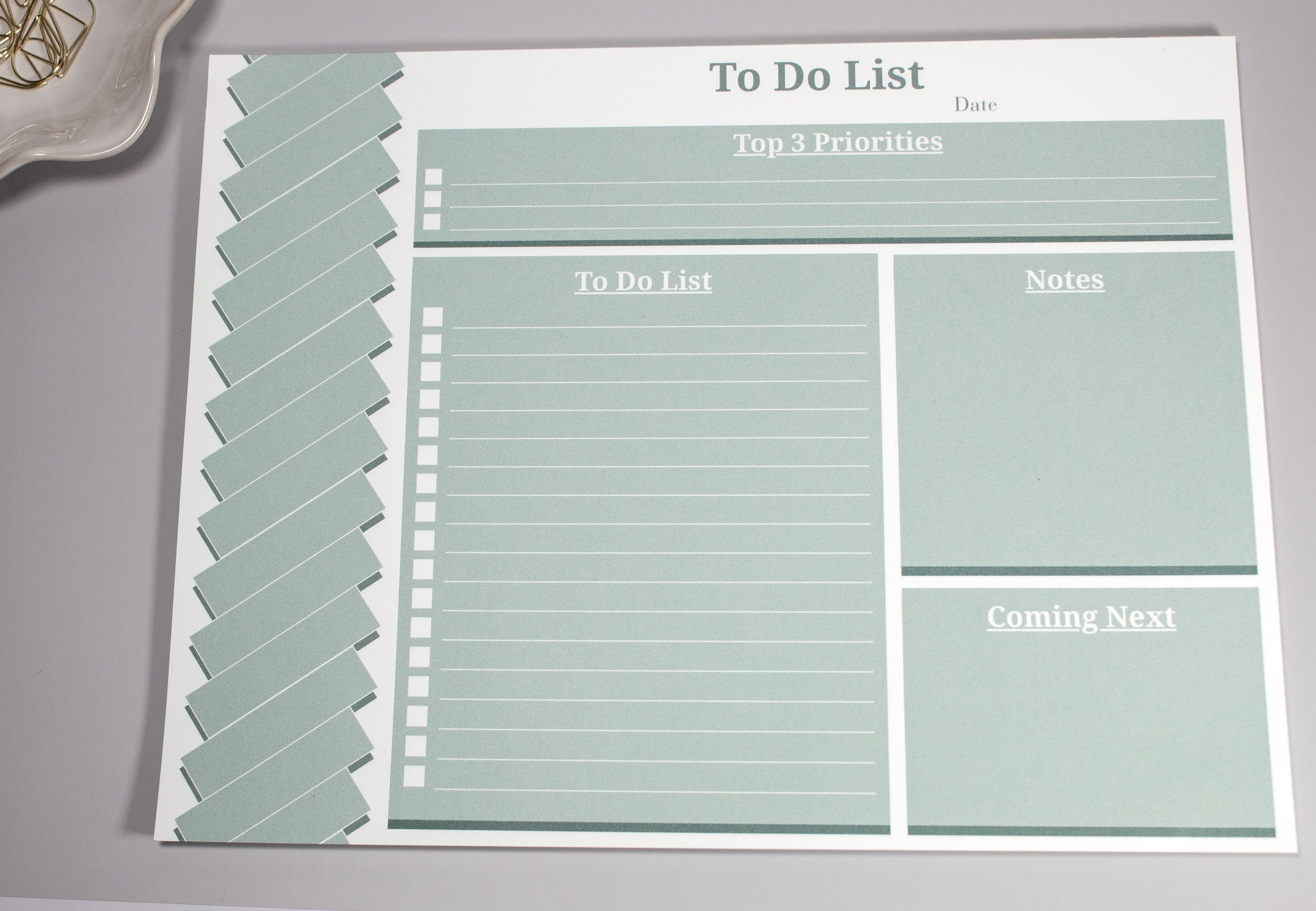 Sage green 8 x 10.5 inch to-do list with a "Top 3 Priorities" box at the top, the "To-do list" section below to the left. On the right of the to-do list is a "Notes" section and a "Coming Next" section below the "Notes Section". There are gold cone-like paperclips at the top sitting inside a flower-like ceramic bowl next to it. 