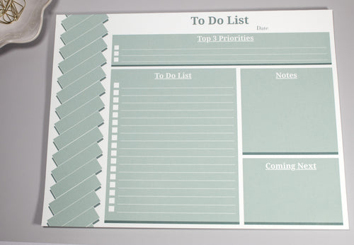 Sage green 8 x 10.5 inch to-do list with a "Top 3 Priorities" box at the top, the "To-do list" section below to the left. On the right of the to-do list is a "Notes" section and a "Coming Next" section below the "Notes Section". There are gold cone-like paperclips at the top sitting inside a flower-like ceramic bowl next to it. 