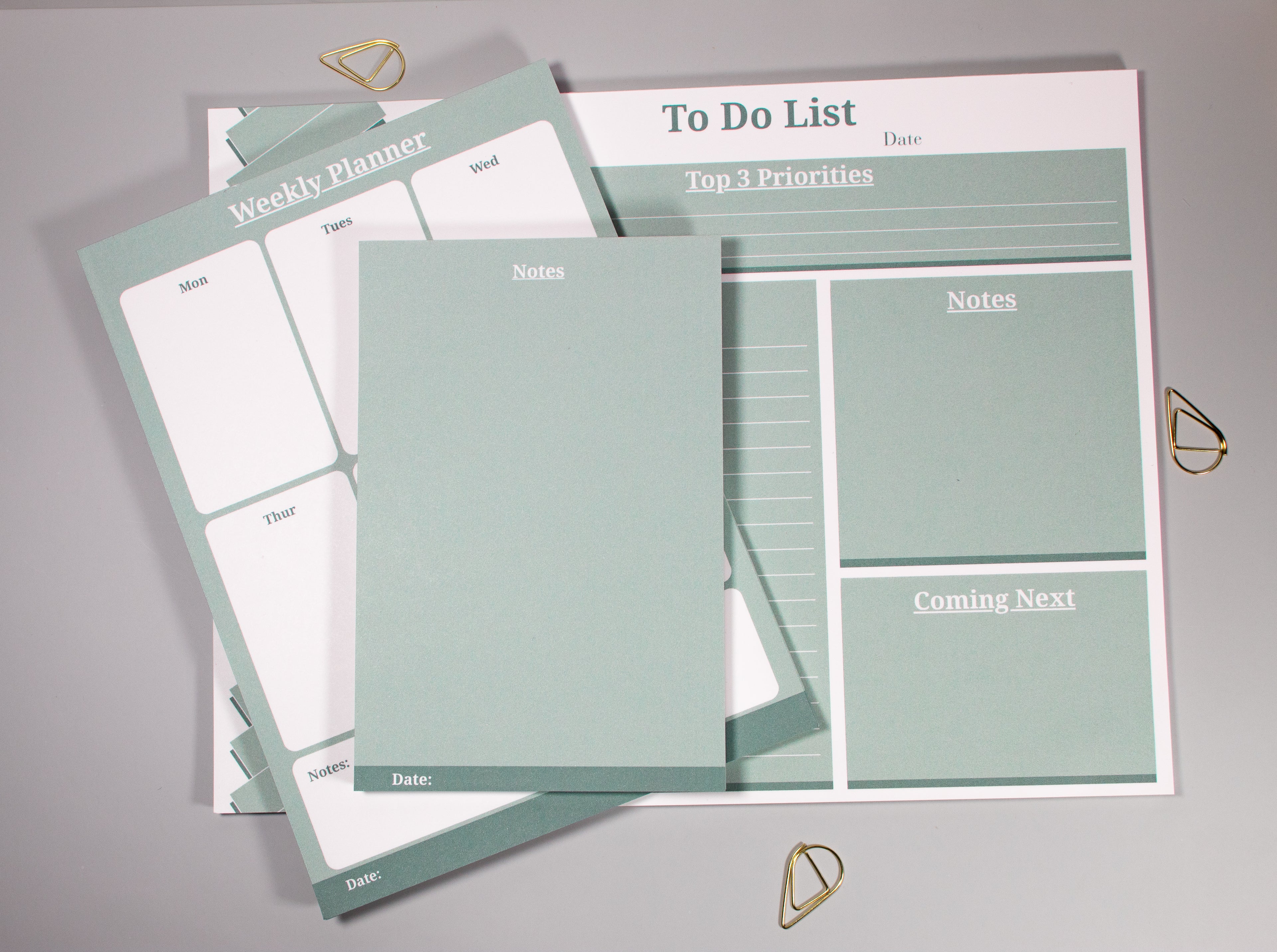 A set of pastel sage green and white stationery items arranged on a light gray surface, including a weekly planner sheet, a to-do list pad, and a notes pad, with small gold decorative cone-like paper clips nearby.