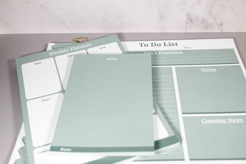 A set of sage green and white paper planner notepads, including a weekly planner, to-do list, notes pad, and priority tracker, arranged on a light gray surface and marble background with a decorative cone-like gold paper clip at the top.
